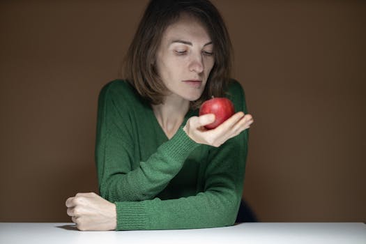 A thoughtful woman in a green sweater examines an apple indoors.
